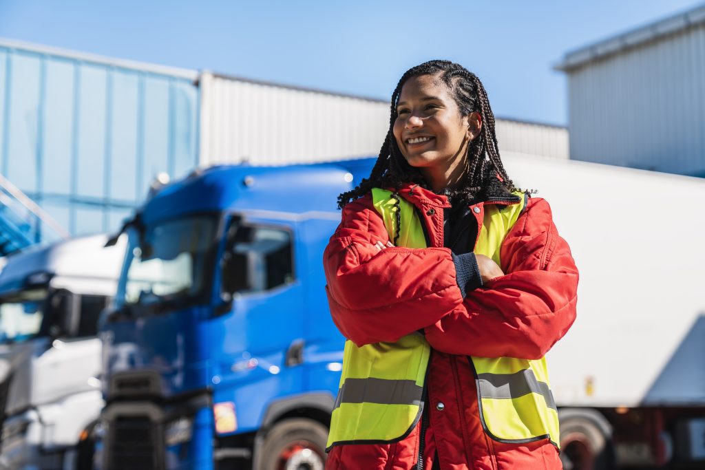 Female professional in reflective vest and warm jacket standing proudly with crossed arms against parked trucks and warehouse